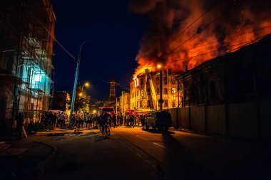 Gece, yanan binanın çatı, itfaiye araçları, itfaiyeciler ve kalabalık insan şehir sokak kazasında izliyor ateş