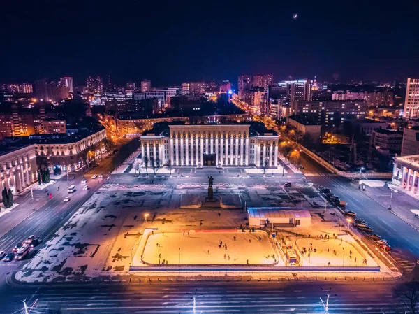 Public ice rink with skating people in city near asphalt road with cars in center or downtown of illuminated winter Voronezh, Russia, aerial view from drone — Stock Photo, Image