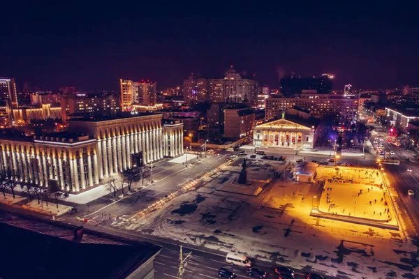 Public ice rink with skating people in city near asphalt road with cars in center or downtown of illuminated winter Voronezh, Russia, aerial view — Stock Photo, Image
