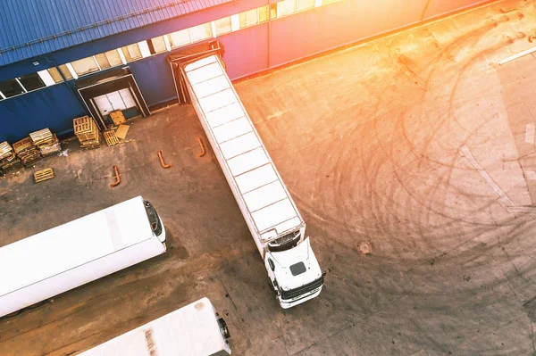 Foto aérea de caminhões de carga em armazém industrial ou centro logístico esperando para carregar mercadorias na luz do pôr do sol, vista superior — Fotografia de Stock