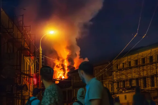 Fogo durante a noite, Telhado do prédio em chamas e pessoas desfocadas em primeiro plano assistindo acidente na rua da cidade — Fotografia de Stock