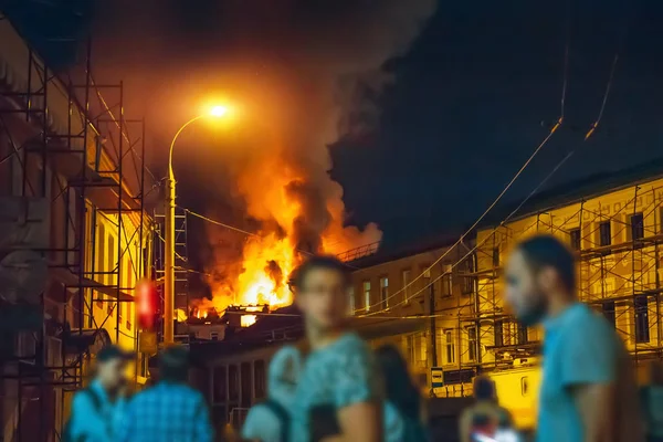 Incendie la nuit, toit du bâtiment en flammes et personnes floues au premier plan regarder accident dans la rue de la ville — Photo