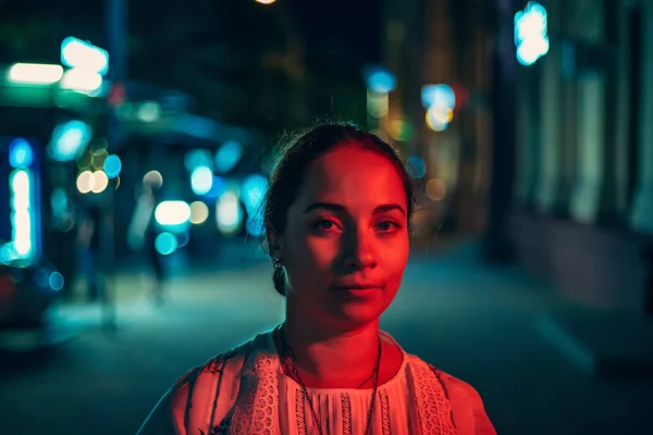 Portrait of young pretty girl looking at camera in red light on her face on night city street with blurred illuminated urban background