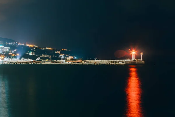 Raios de luz de pesquisa da lâmpada do farol no cais de concreto com reflexão na água à noite — Fotografia de Stock