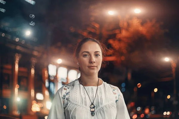 Retrato de mujer hermosa joven mira a la cámara en la calle borrosa iluminada de fondo de la ciudad de la noche — Foto de Stock