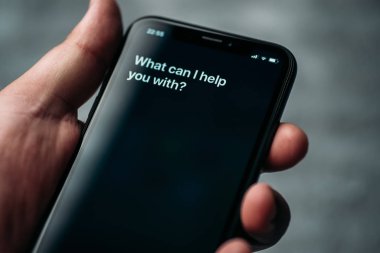Moscow, Russia - Circa August 2019 : Siri - Apple voice digital assistant activated by voice in male hand and text on Iphone screen: What can I help you with? Voice mode of interaction with tphone clipart