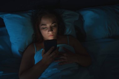 Genç kadın gece uyumak yerine yatakta yatan akıllı telefonu kullanıyor, üstten bakıyor. Uykusuzluk kavramı