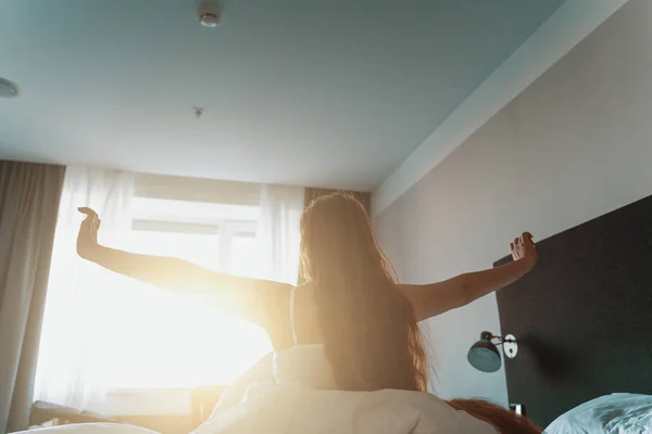Young woman with long hair wakes up, gets up on bed and stretches hands in morning sun light
