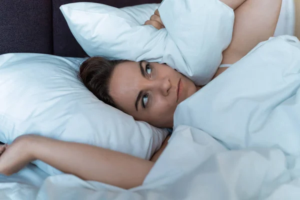Mujer joven estresada no puede dormir debido al ruido de sus vecinos desde arriba, irritada y estresante cubre su cabeza y oídos con almohada —  Fotos de Stock