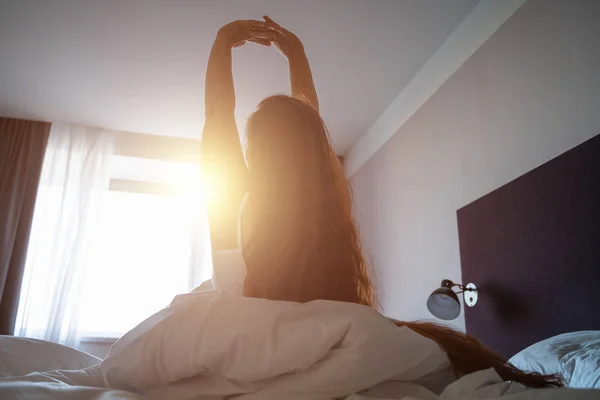 Young woman wakes up, gets up on bed and stretches hands up in morning sunlight — Stock Photo, Image