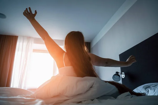 Ung kvinna sträcker händerna i sängen efter vakna i morgon solljus sol, utsikt från ryggen — Stockfoto