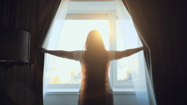 Young woman opens dark curtains and enjoys morning sunlight sun, view from back — Stock Photo, Image