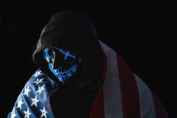 Mann mit Leuchtneonmaske in Kapuze und mit amerikanischer Flagge auf schwarzem Hintergrund. Halloween und Horror-Konzept — Stockfoto