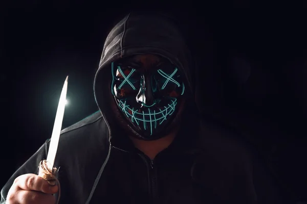 Uomo con illuminazione neon maschera bagliore nel cappuccio e grande coltello in mano su sfondo nero. Halloween e concetto di orrore — Foto Stock