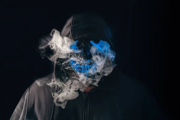 Человек с зажиганием неоновой светящейся маски в капюшоне и паром от маски на черном фоне. Хэллоуин и концепция ужасов — стоковое фото