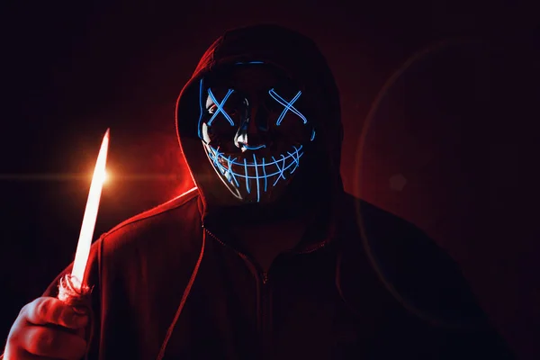 Człowiek z nożem w ręku i zły i przerażające oświetlenie neon blask maska w kapturze na ciemnoczerwonym tle. Halloween i pojęcie horroru — Zdjęcie stockowe