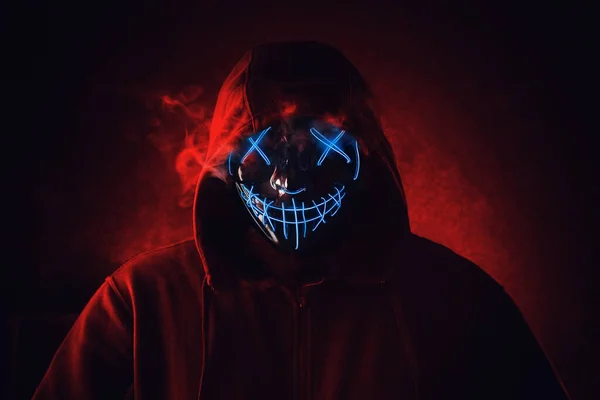 Pria dengan topeng cahaya neon yang menakutkan berkerudung dengan latar belakang merah gelap. Halloween dan konsep horor Stok Lukisan  