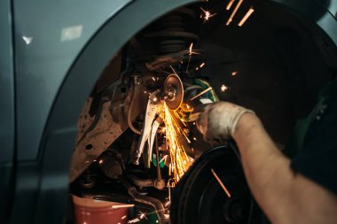 İşçi oto servis istasyonundaki direksiyonun paslı cıvatasını öğütücüyle kesiyor. Kapatın.