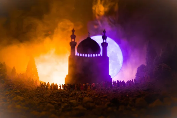 Silhouette of a large crowd of people in forest at night standing against a blurred mosque building with toned light beams on foggy background. Ramadan Kareem background. Praying people concept.