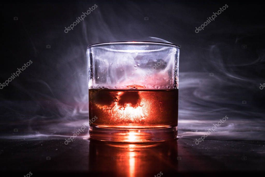 Glass of whiskey and ice on wooden surface with color light and fog on background. Close up. Advertising shot