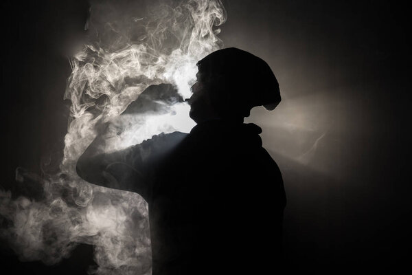 Vaping man holding a mod. A cloud of vapor. dark foggy toned background of clouds of smoke. Vaping an electronic cigarette with a lot of smoke. Vape concept. Selective focus