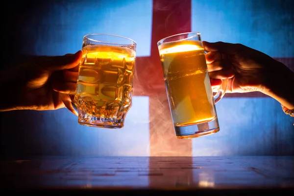 Two friends toasting (clinking) with glasses of light beer at the pub. Beautiful background with blurred view of flag of England. Support your country with beer concept. Selective focus