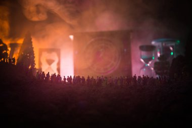 Büyük bir kalabalığa karşı bir tonda ışık ışınları sisli arka plan üzerinde büyük ok saatiyle gece ayakta, ormandaki insan silüeti. Zaman kavramı. Kum saati geçen süre ölçümünü