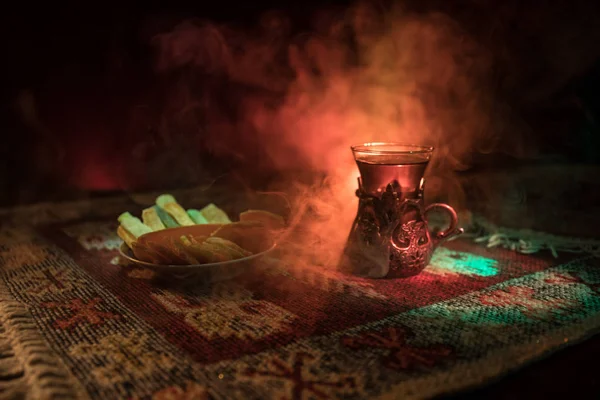 阿拉伯茶在玻璃与东方小吃在地毯上的黑暗背景与灯光和烟雾 东方茶的概念 空的空间 选择性聚焦 — 图库照片