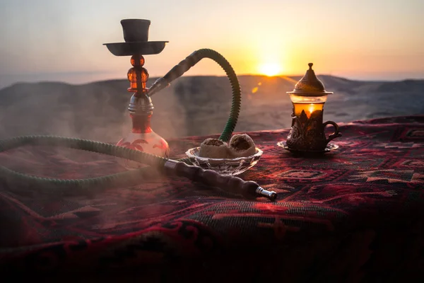 Hookah hot coals on shisha bowl making clouds of steam at desert outdoor. Oriental ornament on the carpet eastern tea ceremony. Stylish oriental shisha on sunset background. Selective focus
