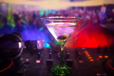 Glass with martini with olive inside on dj controller in night club. Dj Console with club drink at music party in nightclub with disco lights.