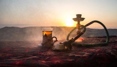 Hookah hot coals on shisha bowl making clouds of steam at desert outdoor. Oriental ornament on the carpet eastern tea ceremony. Stylish oriental shisha on sunset background. Selective focus clipart