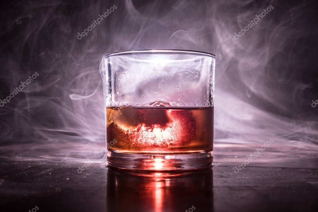 Glass of whiskey and ice on wooden surface with color light and fog on background. Close up. Advertising shot
