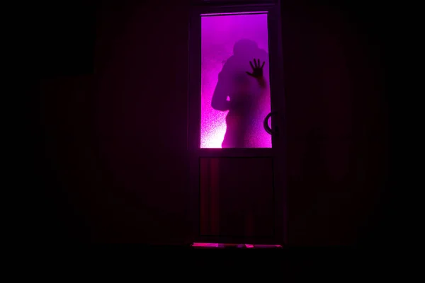 Silhouette of an unknown shadow figure on a door through a closed glass door. The silhouette of a human in front of a window at night. Scary scene halloween concept of blurred silhouette of maniac.