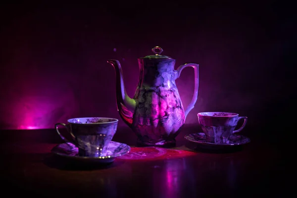 Coffee or tea ceremony conceptual theme. Old vintage ceramic tea or coffee pot with cups on dark toned background with light and smoke. Empty space for text. Selective focus
