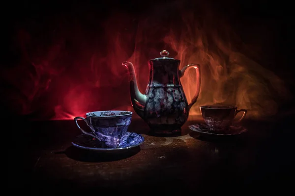 Coffee or tea ceremony conceptual theme. Old vintage ceramic tea or coffee pot with cups on dark toned background with light and smoke. Empty space for text. Selective focus