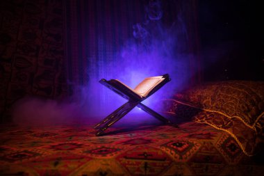 Open holy book of Muslims on stand on eastern carpet with dark toned foggy background. Muslim religion concept. Selective focus. Indoor shot. clipart