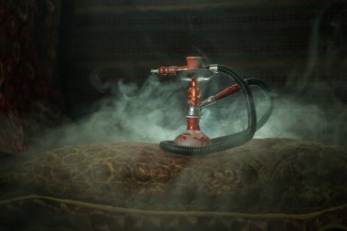 Hookah hot coals on shisha bowl making clouds of steam at Arabian interior. Oriental ornament on the carpet. Stylish oriental shisha with backlight. For Shisha advertisement. Selective focus clipart
