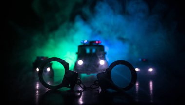 Police raid at night and you are under arrest concept. Silhouette of handcuffs with police car on backside. Image with the flashing red and blue police lights at foggy background. Slider shot clipart