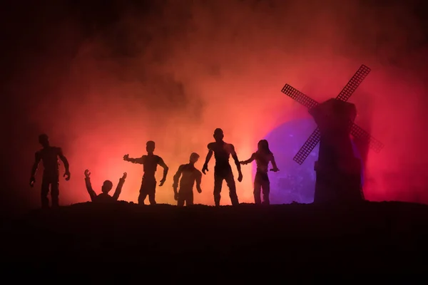 Silhouettes crowd of hungry zombies and old windmill on hill against dark foggy toned sky. Silhouettes of scary zombies walking at night. Horror Concept