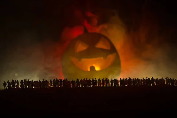 Halloween concept. Blurred silhouette of giant Jack-o-lantern pumpkin with scary smiling face behind crowd at night. People looks at a big pumpkin at night. Selective focus.