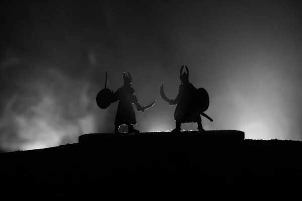 Silhouette of two medieval warriors in duel. Picture with two fighter with sword with dark toned foggy background. Selective focus.