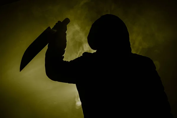 A dangerous hooded man standing in the dark and holding a knife. Face can not be seen. Committing a crime concept