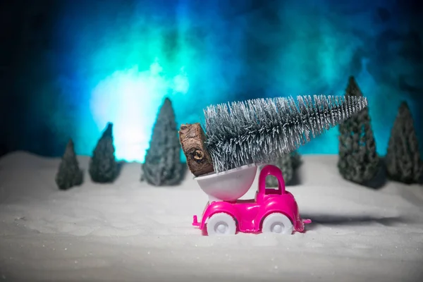 Christmas or New Year concept. Toy car carrying a Christmas tree through the forest in snowfall. Holiday decorated background. Selective focus