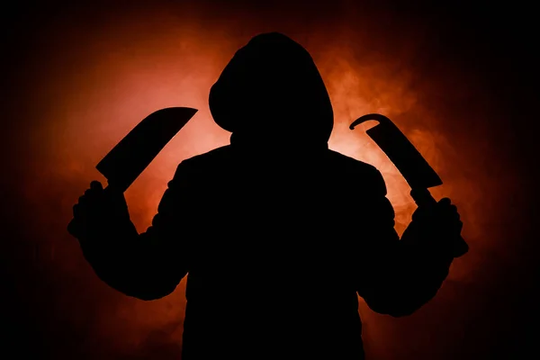 A dangerous hooded man standing in the dark and holding big knives. Face can not be seen. Committing a crime concept