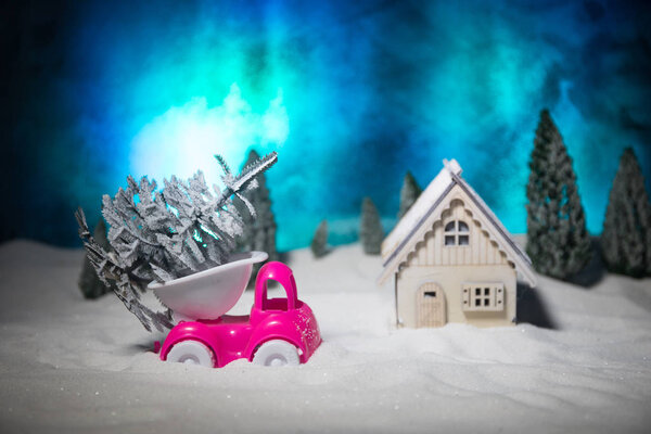 Концепция Рождества или Нового года. Игрушечный автомобиль, несущий елку через лес в снегопад. Праздничный декорированный фон. Селективный фокус
