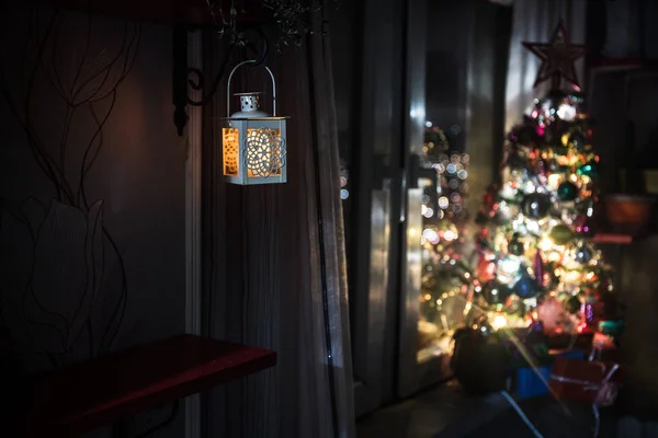 desk of free space and lamp with xmas tree in home . Christmas lantern in selective focus near window with holiday tree full of colored toys and lights. Night scene