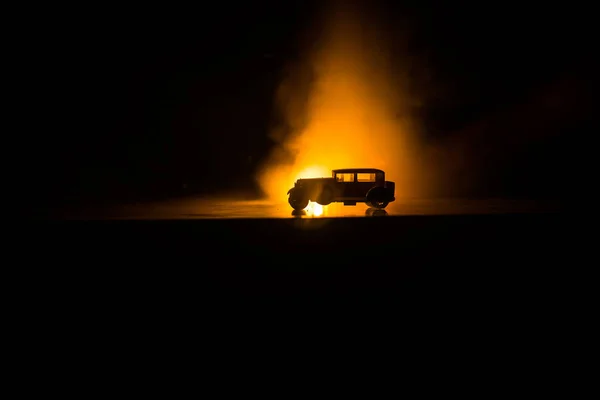 Silhouette of old vintage car in dark foggy toned background with glowing lights in low light. Selective focus