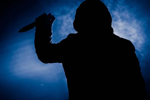 A dangerous hooded man standing in the dark and holding a knife. Face can not be seen. Committing a crime concept