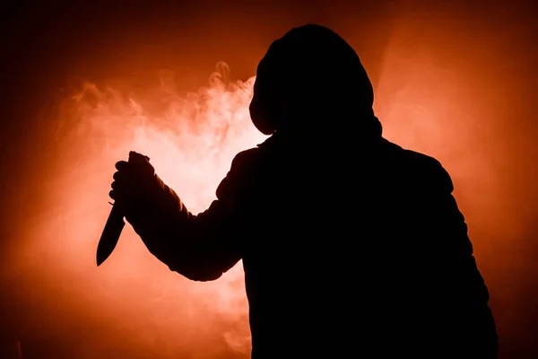 A dangerous hooded man standing in the dark and holding a knife. Face can not be seen. Committing a crime concept