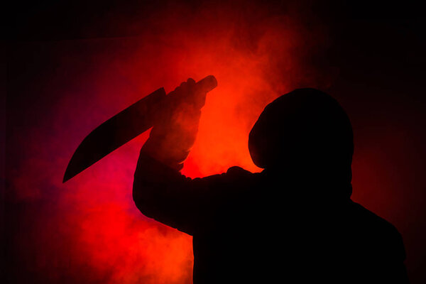 A dangerous hooded man standing in the dark and holding a knife. Face can not be seen. Committing a crime concept